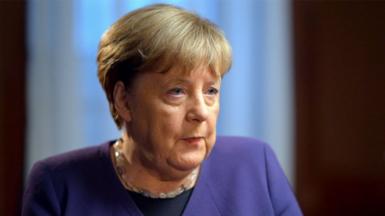 Head shot of Angela Merkel being interviewed. She is looking to the right and is wearing a purple jacket and a necklace