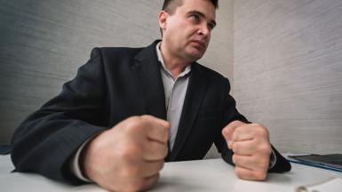 Man in suit looks angry and clenches his fist on the desk in front of him