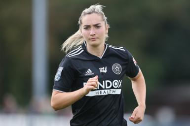 A young woman with long blonde hair tied back wears a black football shirt as she jogs forwards, with a look of concentration on her face. The shirt has various logos and a Sheffield United team crest printed on it.