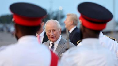 King Charles, in focus, stands between two out-of-focus soldiers wearing white uniforms and black top hats with red bases.