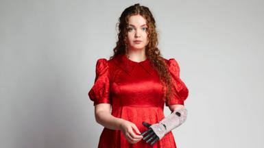 Gracie McGonigal has long auburn curly hair and wearing a red dress. She is wearing a prosthetic hand which is glittery and has black coloured fingers. 
