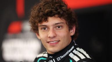 A young motorsport driver with curly brown hair, dressed in a sleek black racing suit accented with white and teal details. Sponsor logos are visible across the suit, including one on the collar. 