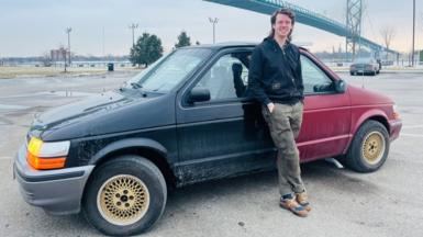 A man is in a car park leaning on a car made of two front ends - one red and one black. A bridge is in the background. 