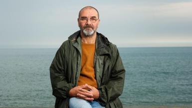 Andrew Malkinson sits with his hands clasped together. He wears jeans, an orange jumper and green coat and looks straight at the camera, unsmiling. He is pictured against a backdrop of the sea. 