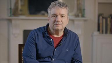 Dr Ittai Gradel looking to camera in the centre of the image. In blue shirt and maroon top underneath. Behind him books and fire place