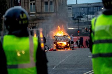 Police in riot gear look on with a burning car in the road in Sunderland