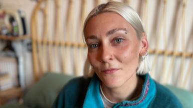 Elle Glenny, who has bobbed blond hair, looking directly at the camera and wearing a turquoise fleece. There is a bed frame in the background.