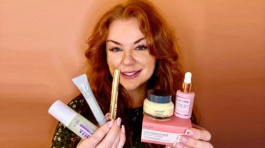 A ginger woman is holding up her favourite skin care to the camera. In one hand she has three tube like products and in the other she has another three products, one if in a box another in a tub and a third in a bottle