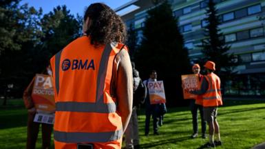 A person with their back to the camera wears a hi vis orange vest with the letters 'BMA' on the back, watching as resident doctors stand with placards while picketing outside Manchester university hospital on Tuesday.