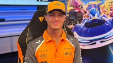 Lando Norris wearing an orange cap, and an orange polo shirt. He is sitting in front of a poster which includes sonic the hedgehog in a blue car.