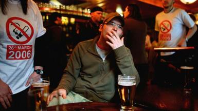 A man sitting at a table in a bar smokes a cigarette with a pint in front of him. Behind him, people are wearing t-shirts with a smoking ban symbol and the date of 26 March 2006