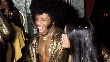 Sly Stone and his newlywed wife Kathy Silva grin and laugh, dressed in gold. She has confetti scattered through her hair.