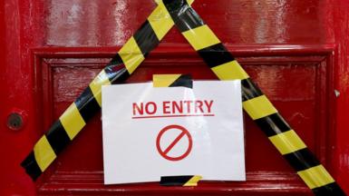 A red door has yellow warning tape across it in the shape of an X. In the middle of the door a laminated A4 sign reads no entry in red writing.