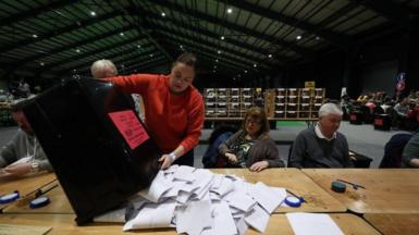 A woman wearing a red top empties a large black bin containing ballot papers onto a desk. To her right, a woman and a man both sit at the desk ready to count. They are in a large hall. In the background a number of other people sat at desks can be seen.