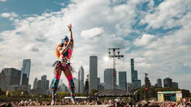 Chappell Roan wears a pink leotard emblazoned with stars, thigh-high boots with stars and a super hero mask. She is standing on a stage with one hand raised in the air.
