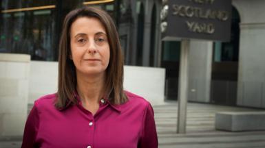 Lucy Manning, a woman in a pink blouse with brown hair, stands in front of a building looking at the camera. There is a sign that says "New Scotland Yard".