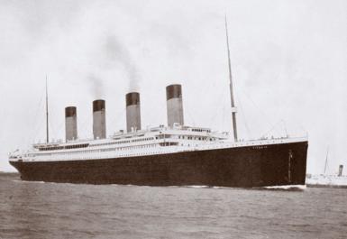 A black and white photo of RMS Titanic, a large ocean liner with four funnels, a black hull and a white upper deck. There are masts at the front and rear.