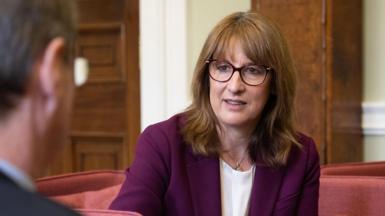A close up image of Rachel Reeves wearing glasses and a purple suit while sitting on a sofa as she talks to Sir Charles Roxburgh (out of frame) in London on Monday.