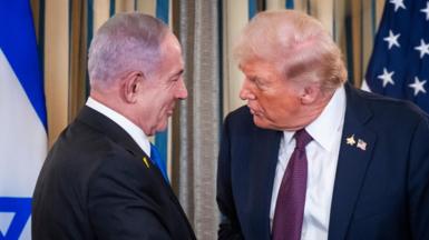 US President Donald Trump (R) shakes hands with Israeli Prime Minister Benjamin Netanyahu (L), with Israeli and American flags behind them. Both men are wearing dark suits and white shirts.  