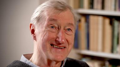 Julian Barnes smiling, top of checked shirt collar & dark top. Book shelf behind him.