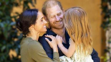 Duke and Duchess of Sussex in casual outfits embracing and holding Lilibet in a summer dress, she has bare feet and her back is to the camera; she has long, red hair
