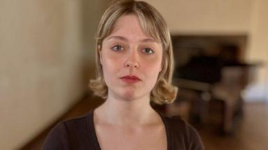 Lizzie Blissett looks directly at the camera. She is wearing a black scoop-neck dress and has her blonde hair in a bob. Behind her, blurred, is a piano.