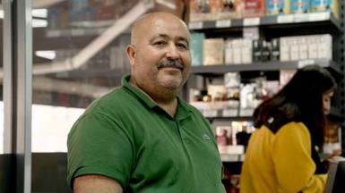 Erol Kaya in a shop wearing a green shirt facing the camera, behind is a woman in a yellow shirt looking down, with shelves of products in the background.
