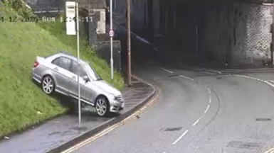 Image take from a CCTV video shows a silver car bouncing onto a stretch of pavement heading towards a main road from a grass hill.