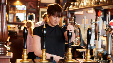 Woman with dark brown hair wearing a black short-sleeve top pours pint or beer with pump behind a bar in a pub