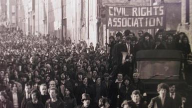 A black and white photograph of hundreds of people marching along a street in Londonderry on 30 January 1972.
On the right hand side of the image is a truck, with people standing on the back holding a large sign which says Civil Rights Assocation.