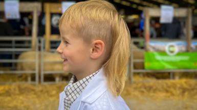 Boy in a sheep shed wearing a white coat for showing and with a long blonde mullet 