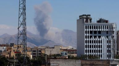 Smoke rises in the mountains in the distance as we look at building tops from far away in Iran as well as a metal tower.
