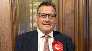 Karl Turner the succesful Labour candidate for Hull East pictured on election night in 2024 wearing a dark suit jacket, white shirt, and patterned pink tie, standing indoors against a wooden paneled wall. A large red rosette with the word ‘Labour’ and smaller text is pinned to the left side of the jacket. 