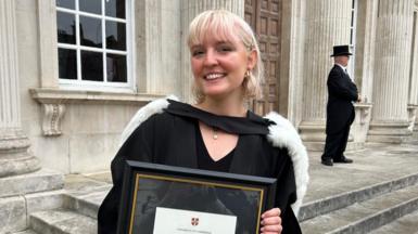 Lucy Gabb, with short blonde hair, smiling while wearing her graduation gown and holding her degree certificate.
