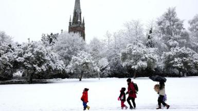 Snowy scene with a park area and a church in the background.  Trees covered in snow and a group of people walking in the snow.
