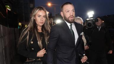 Conor McGregor wears a black suit, white shirt and green tie. He is holding his wife hand who has blonde hair and wears a black suit with black top, holding a balck and gold handbag 