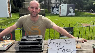 Patrick, a man with a shaved head and stubbled facial hair, smiles sitting behind his typewriter on a wooden picnic bench in Botanic. In front of his typewriter is a handmade sign on a piece of folded white paper saying "Free typewriter poems" in Botanic Gardens in Belfast.