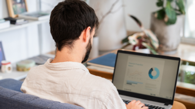 Young man with dark hair and beard sitting on a blue couch in beige shirt with his back to the camera, looking at a laptop screen showing a financial report with graphics