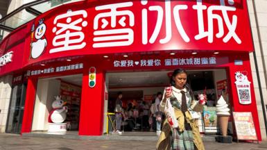 A shop of Mixue, a Chinese bubble tea brand in Chongqing, China in April 2026. In the foreground a girl walks away from the shop holding two sundaes and a drink.