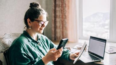 Woman sits at a window seat holding a phone and a pad with a computer on a table in front of her.