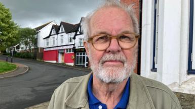 Photo of Kjell-Åke Andersson who is a man with grey hair and a grey beard. He's  wearing brown rimmed glasses and wearing a blue polo tshirt and faded green jacket. Behind him over his right shoulder is a road and some terraced buildings including a rugby club entrance. The sky has grey clouds and there is also a patch of grass and part of a green tree visible to the left of the frame.