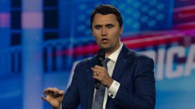 Charlie Kirk holds a microphone in one hand and gestures with the other. He's wearing a blue suit and tie and standing in front of a red, white and blue background.