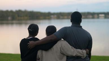 Marven, Rochelle and Guerline, pictured from behind, with their arms around each other looking out over a lake. Marwen is wearing a grey, collared T-shirt and is much taller than the two women.