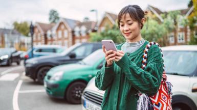 A young woman, dressed in a green cable-knit cardigan uses her phone in a car park, five cars are parked behind her, a silver, green, black, blue and black car. 