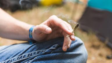 A close-up stock photo that shows the knee and hand of a man wearing jeans sitting cross legged.  He's holding what appears to be a half-smoked cannabis joint.