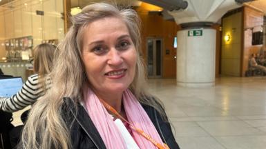 Katalin Kaszas smiling while seated indoors, wearing a pink scarf and black jacket.