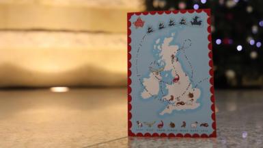 A map of the United Kingdom with a Christmas-themed design, showing a trail of Santa’s sleigh and various holiday icons such as reindeer, robins, and other animals, held in front of a decorated Christmas tree with ornaments