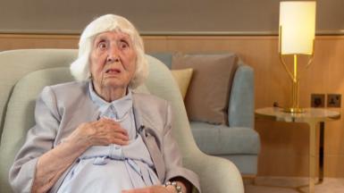 Betty Brown wearing a blue dress, grey jacket and pearls sits in a green armchair. In the background is a lamp on a table.