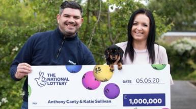 Anthony Canty is wearing a navy quarter zip jumper. He has dark hair and is holding a large cheque for £1m outdoors. Also holding the cheque, and a small black dog, is his partner, Katie Sullivan. She has long dark hair. Both are smiling.