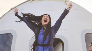 A Vietnamese woman in a shiny cobalt blue jumpsuit is pictured with her fists in the air and mouth open wide as she cheers whilst leaving the Blue Origin rocket, pictured behind her. The rocket is white, with two window panels either side of the door. Another woman smiles up at the astronaut.